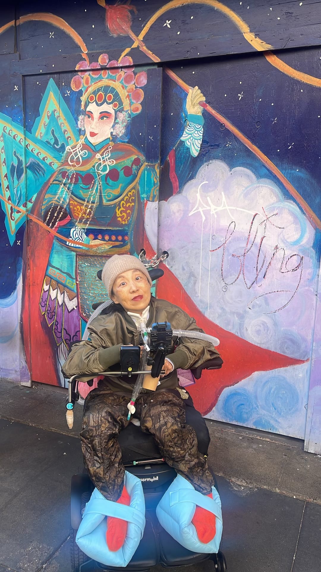 a photo of me, an Asian American woman in a wheelchair with a tracheostomy and a tube attached to a ventilator. I’m wearing a beanie, olive green jacket and camouflage pants. Behind me is a mural featuring a Chinese opera singer 