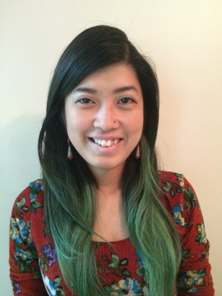 The author is smiling, wearing a floral red dress with long black and green hair. Photo credit: Joe Martello