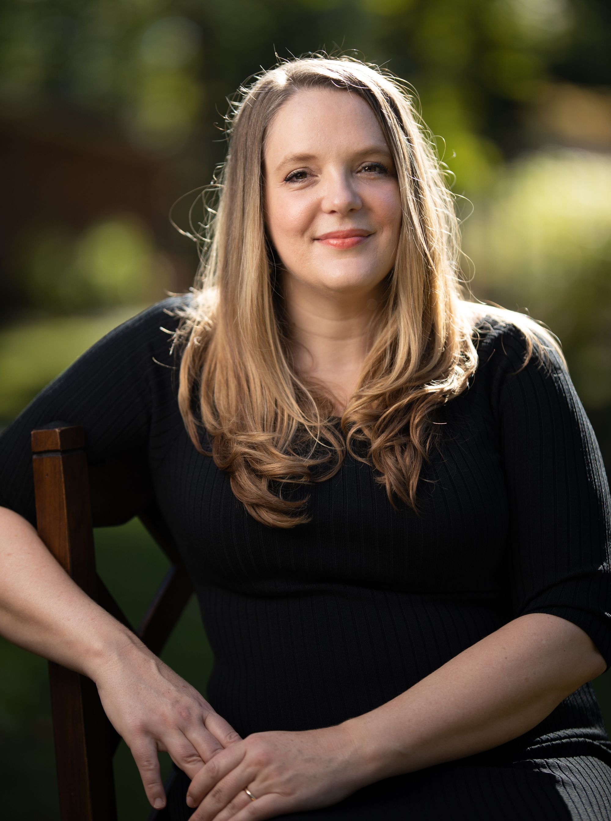 Dr. Cassandra Quave sitting outside in a wooden chair against a blurred green background. She is wearing a black dress, her straight blonde hair is down, and she is slightly smiling at the camera. Photo credit Kemi Griffin