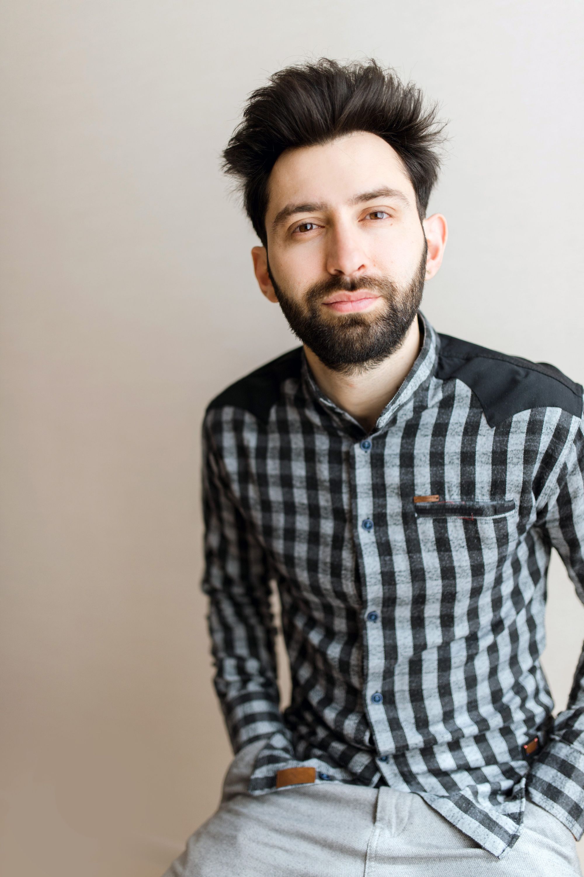 The author, wearing a black and grey checkered shirt and grey pants, sits with hands in pockets against a light beige background. 