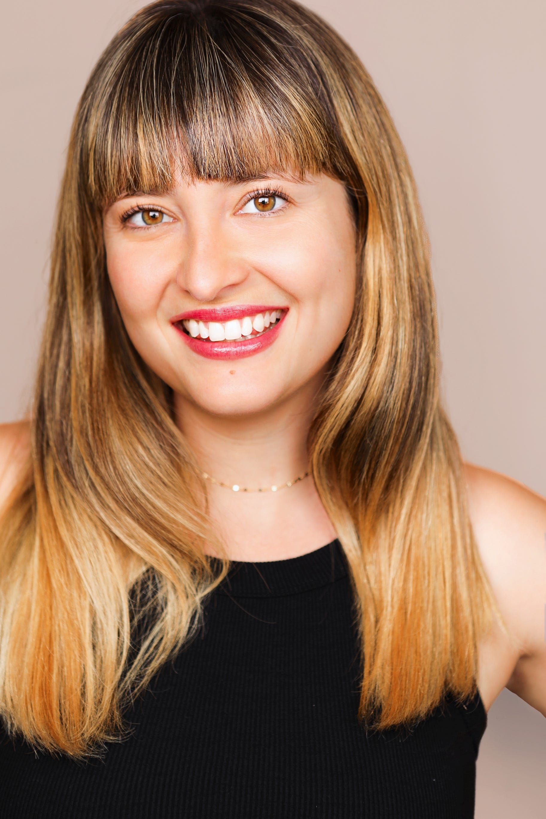 White woman with brunette, chest length, straight hair and bangs smiling and looking directly into camera wearing a black tank top and a gold necklace. Photographed from the waist up. Photo credit: Alexis Dickey. 