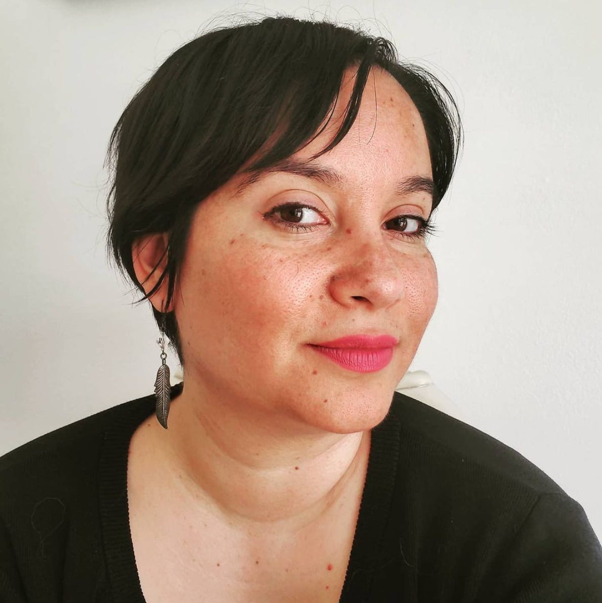 Shayda, a Middle Eastern femme wearing bright pink lipstick. She smiles at the camera. She has short, wispy hair, a long silver feather earring, and is wearing a black shirt.  