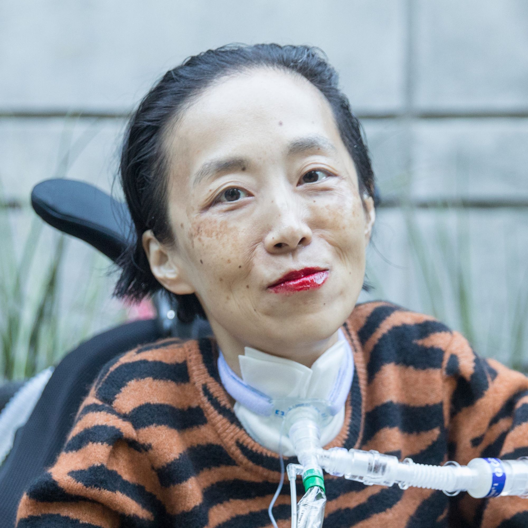 Photo of Alice Wong, an Asian American disabled woman in a power chair. She is wearing an orange and black tiger-striped sweater, black pants, a bold red lip color and a trach at her neck. In the background is a gray cement wall with greenery. Photo credit: Eddie Hernandez Photography.