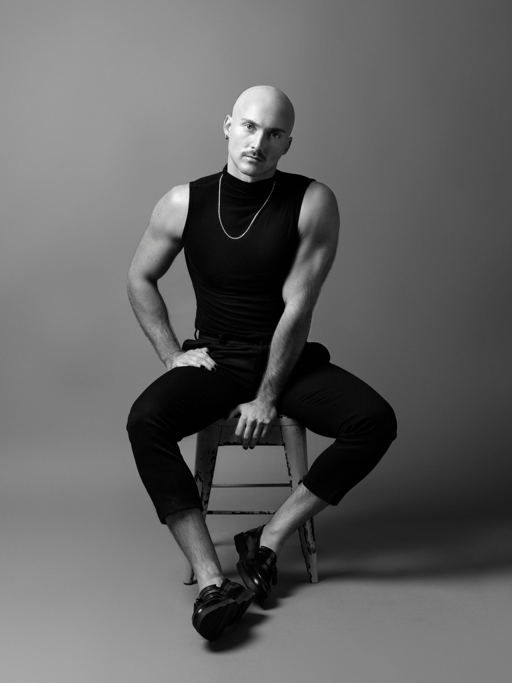 Seated on a stool, Logan is a white person with a mustache and shaved head, wearing a black sleeveless top, silver chain necklace, black pants, and black shoes. They stare directly into the camera without smiling. Photo credit: Hampus Danielsson