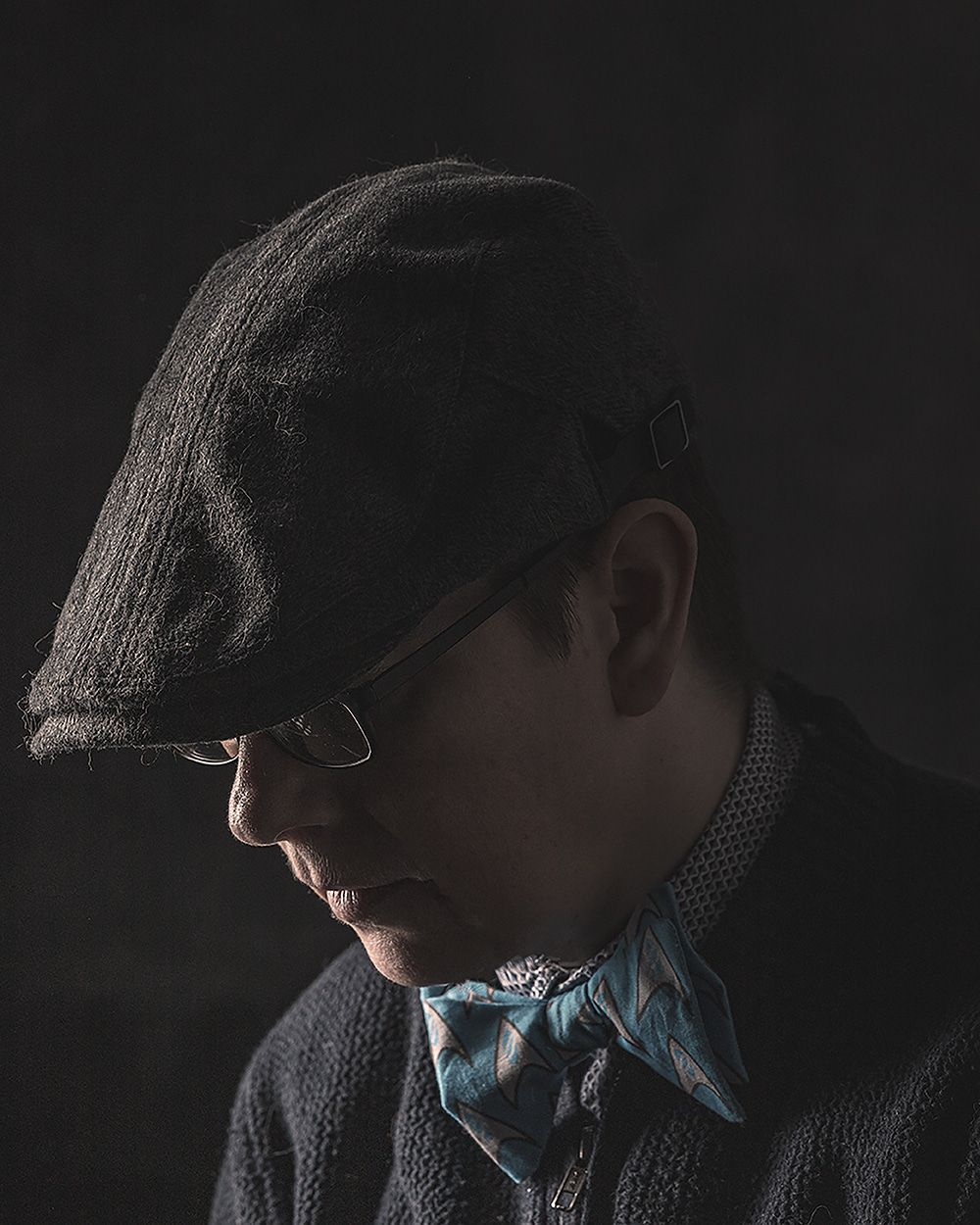 Shoulder-up portrait shot, Jules is a white person with brown hair, wearing thin black glasses, a black newsboy cap, blue and grey Star Trek bowtie, grey patterned collared shirt, and knitted navy-blue zip up cardigan. He is looking down without smiling. Photo credit: Jules Sherred.
